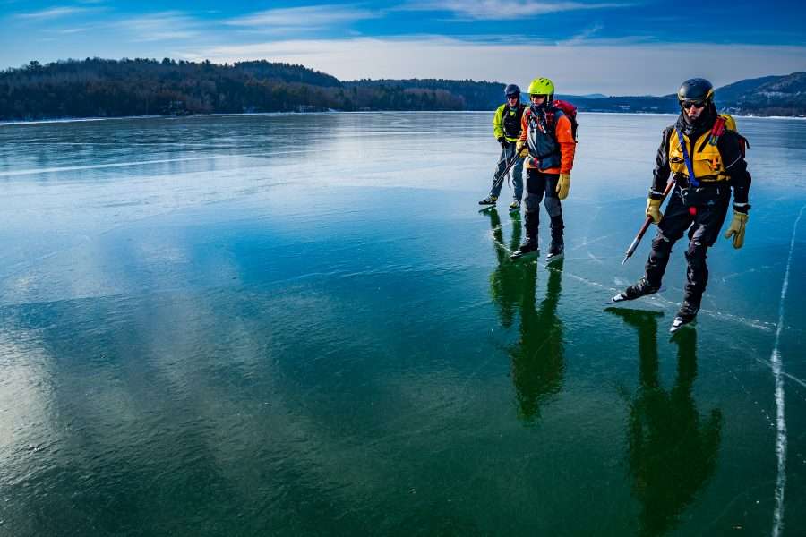 Dan Spada, John Rosenthal, and Dave Phillips are pictured skating across a blue and icy Lake Champlain off Fort Ticonderoga in helmets, vests and other gear. They practice wild ice skating and climate change is altering how often they can skate outside.