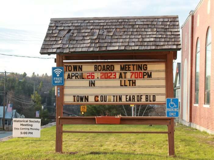 A small a-frame sign with a shingled roof shows details of a town board meeting outside of Long Lake Town Hall.
