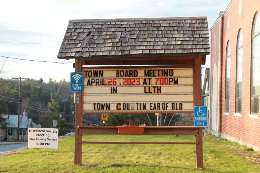 A small a-frame sign with a shingled roof shows details of a town board meeting outside of Long Lake Town Hall.
