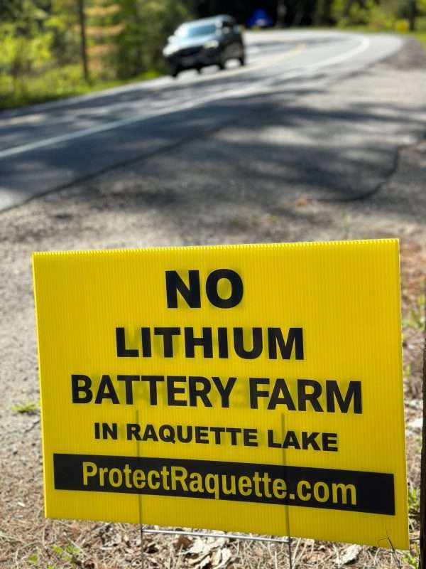 A yellow sign saying "No lithium battery farm in Raquette Lake" sits by the road.
