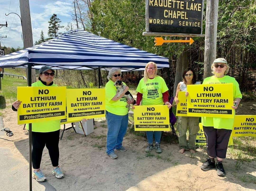 A group of people stand outside with signs that say "No lithium battery farm in Raquette Lake."