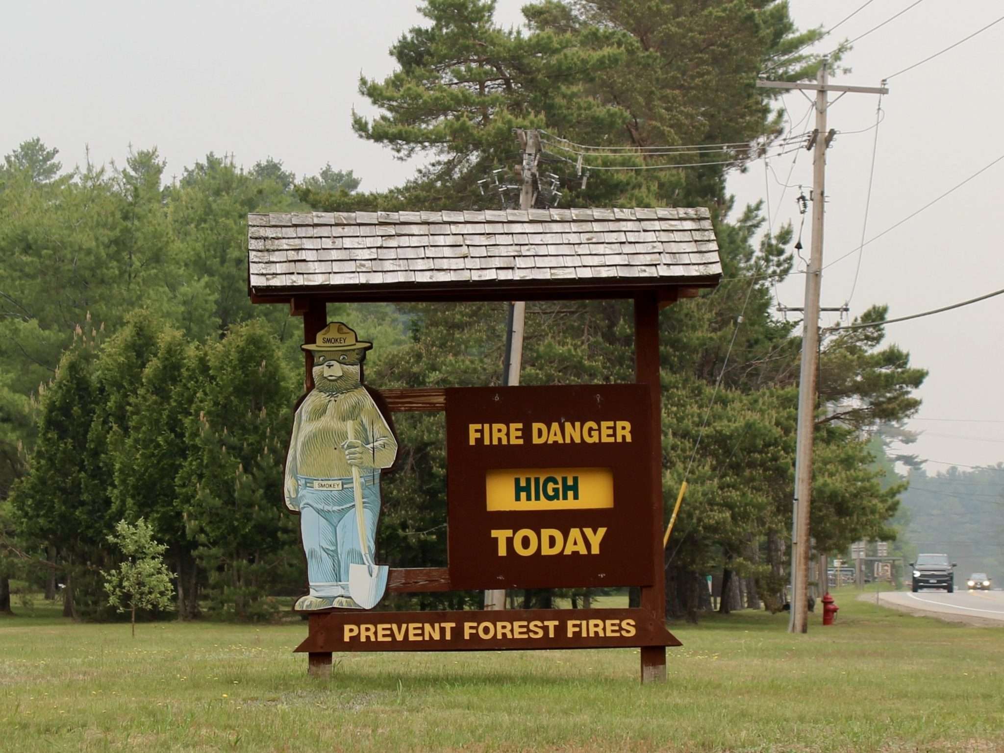 A wooden sign with Smokey Bear in front of the Department of Environmental Conservation shows that fire danger is "high" today. Hazy skies from smoke are behind it.