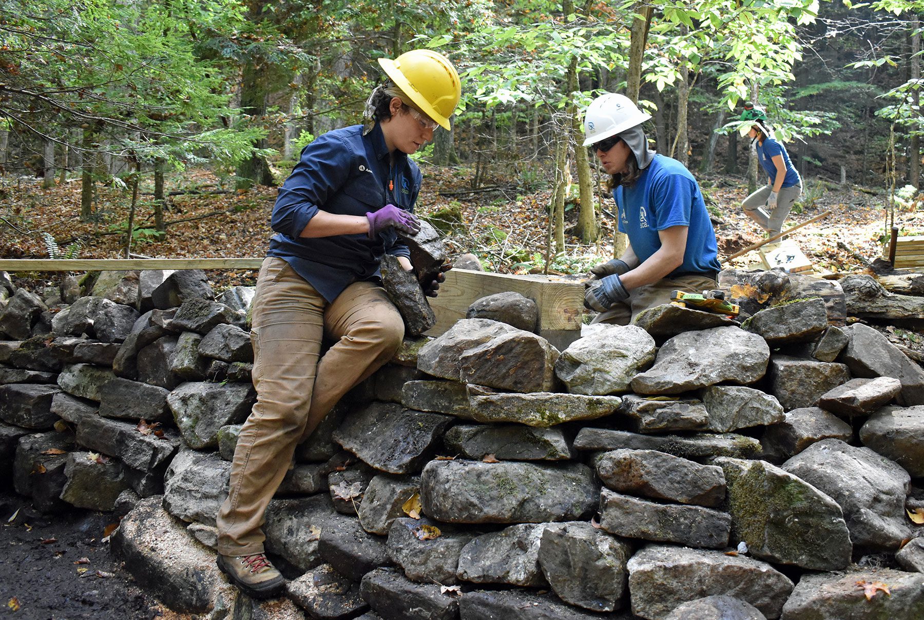 Two people wearing blue shirts and yellow hard hats are working on a stone wall