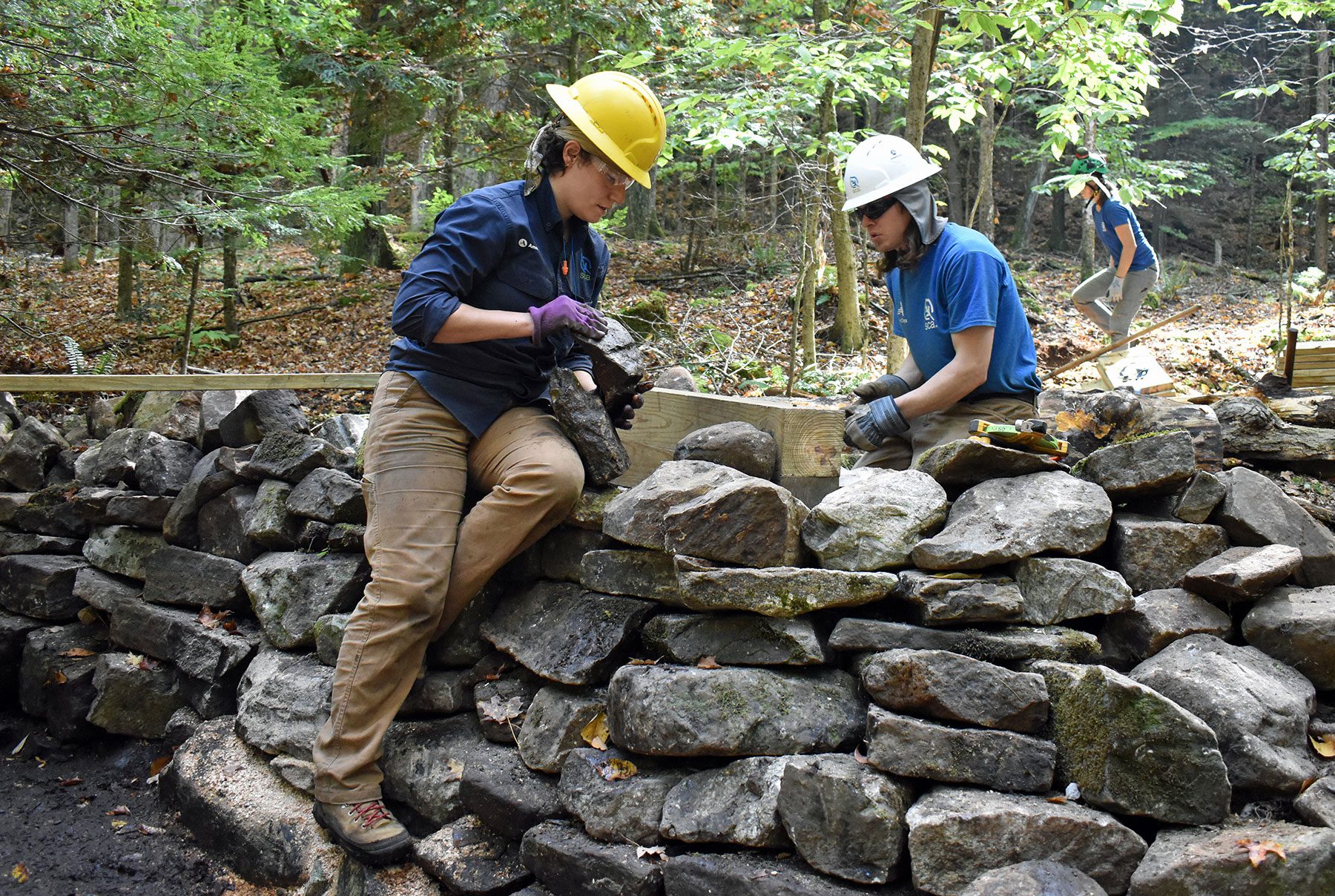 Two people wearing blue shirts and yellow hard hats working on a stone wall