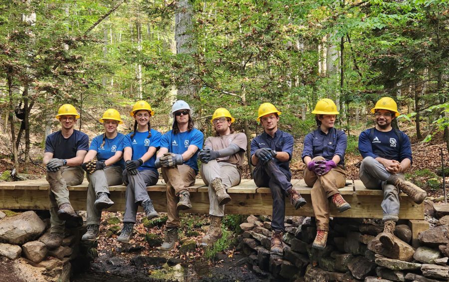 group of student conservation association student volunteers wearing blue shirts and yellow hard hats sitting on a bridge they built