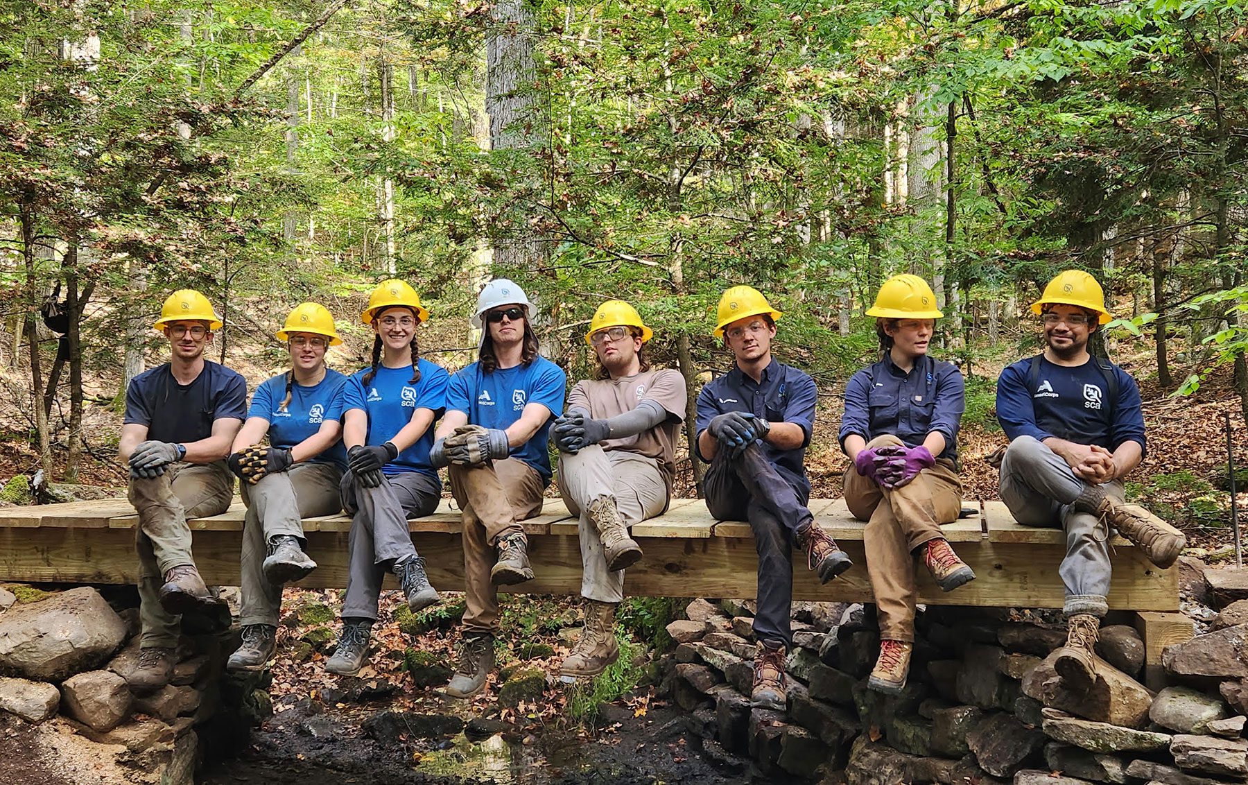 group of SCA student volunteers wearing blue shirts and yellow hard hats sitting on a bridge