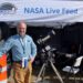 smiling man with telescope and NASA live feed tent behind him