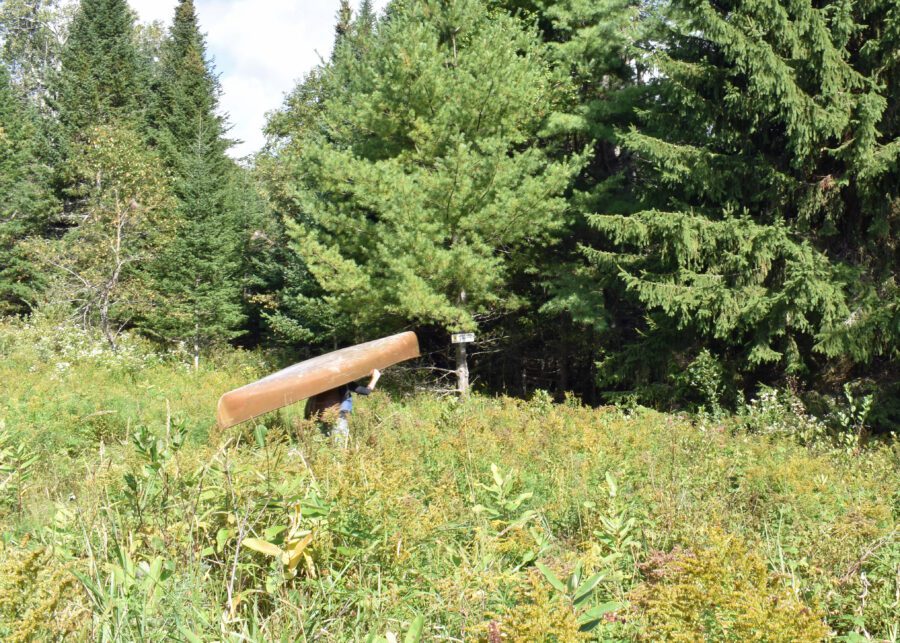 Man carries brown canoe through grassy field to Mud Pond on the former Cedarlands Scout Reservation