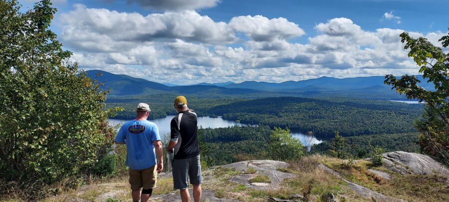 Two men look out at mountains and lakes from a mountain peak.
