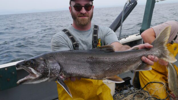 Man in yellow waders on a boat holding a large lake trout in front of him