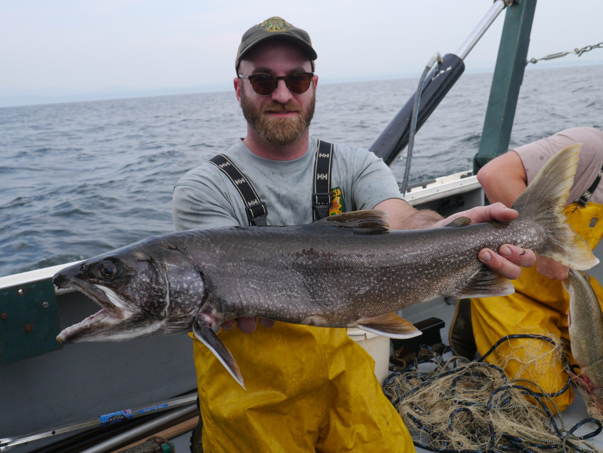 Man in yellow waders on a boat holding a large lake trout in front of him
