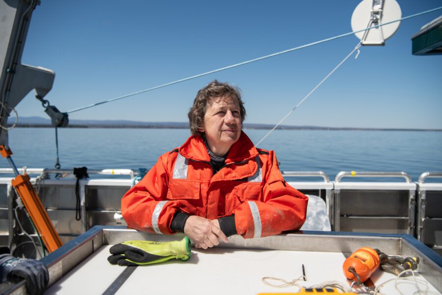 A woman in orange nautical life vest standing on deck of a boat with lake in the background