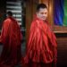 man draped in shiny red fabric with his reflection in a mirror and pride flag