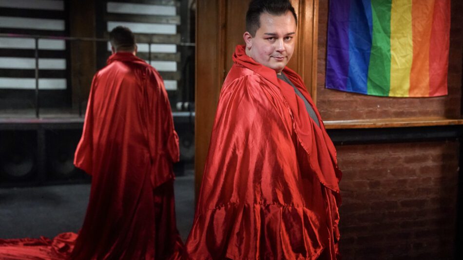 man draped in shiny red fabric with his reflection in a mirror and pride flag