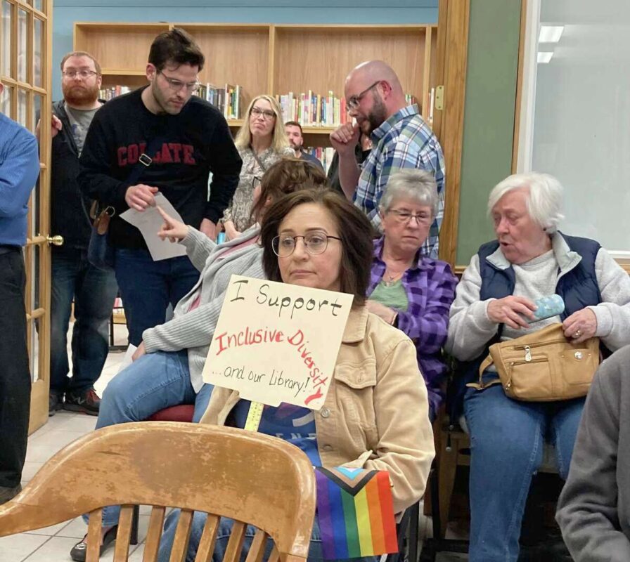 a woman hold a sign in the foreground in support of a drag queen story hour, during a meeting at the library
