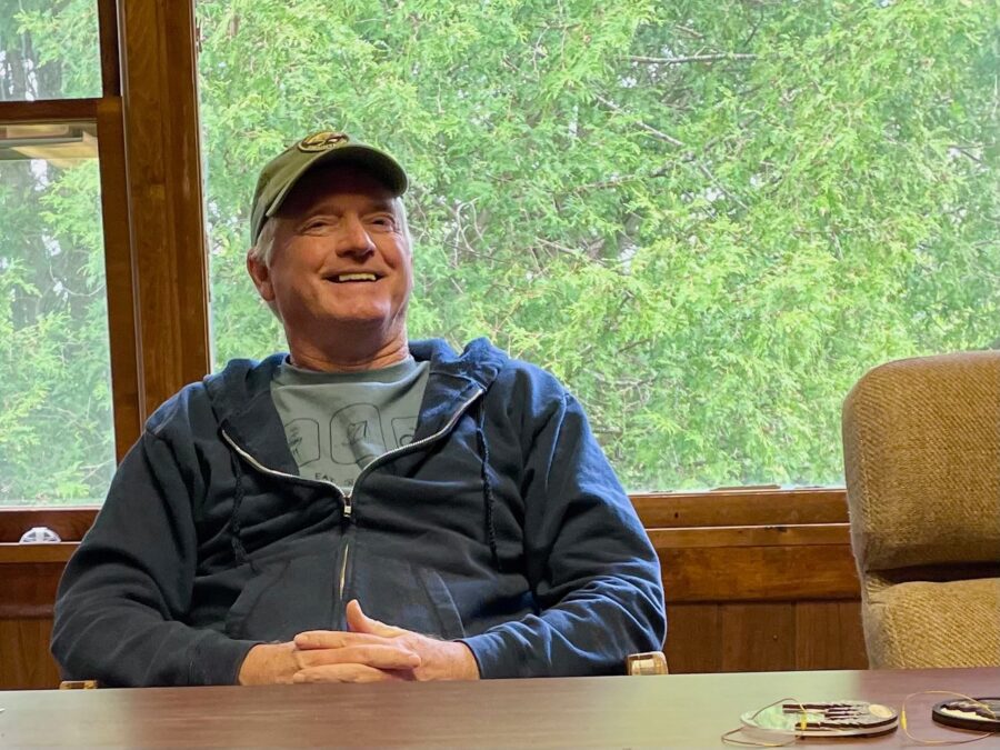 man at a desk in a blue sweatshirt and green cap smiling