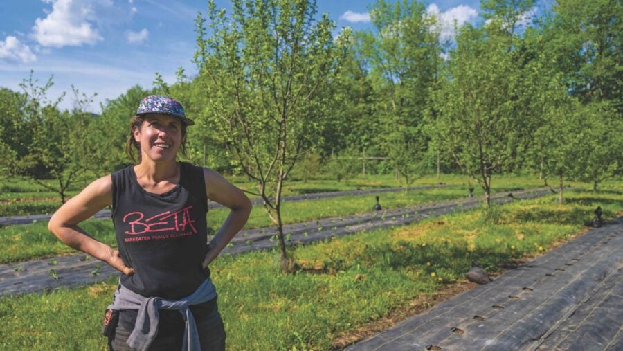 A woman farmer stands in front of a tree and crops