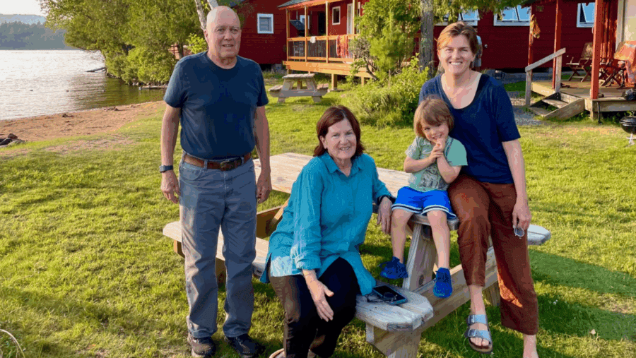 A family including an older man in a blue tee shirt and blue jeans, an older middle aged woman in a blue button down shirt and black slacks and a younger woman with a child in her lap sitting on a picnic table on grass outside of a cabin
