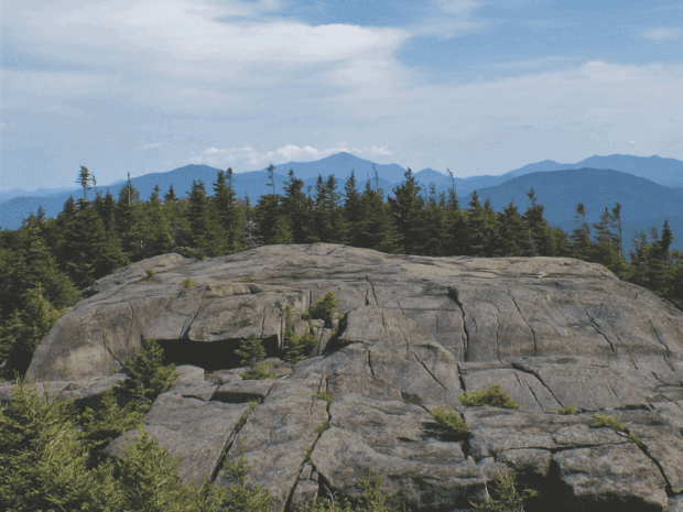 View from bald face mountain top over tree lines looking toward blue Adirondack mountain vista in the distance.
