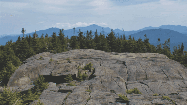View from bald face mountain top over tree lines looking toward blue Adirondack mountain vista in the distance.