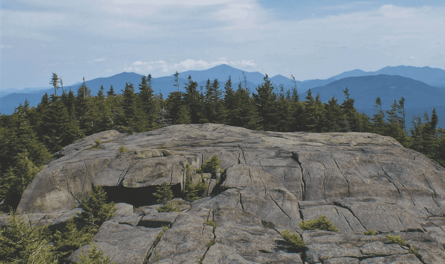 View from bald face mountain top over tree lines looking toward blue Adirondack mountain vista in the distance.
