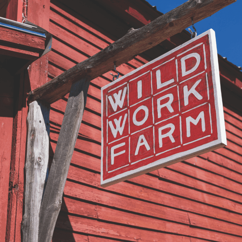 Square sign hanging in front of a red building that says Wild Work Farm with white letters on red background