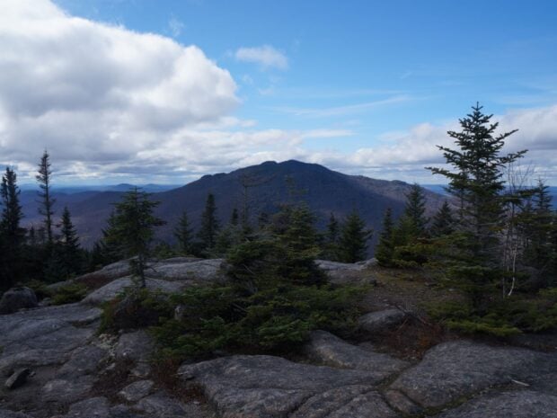 Rock face mountain summit looking toward alpine trees and another small mountain in distance