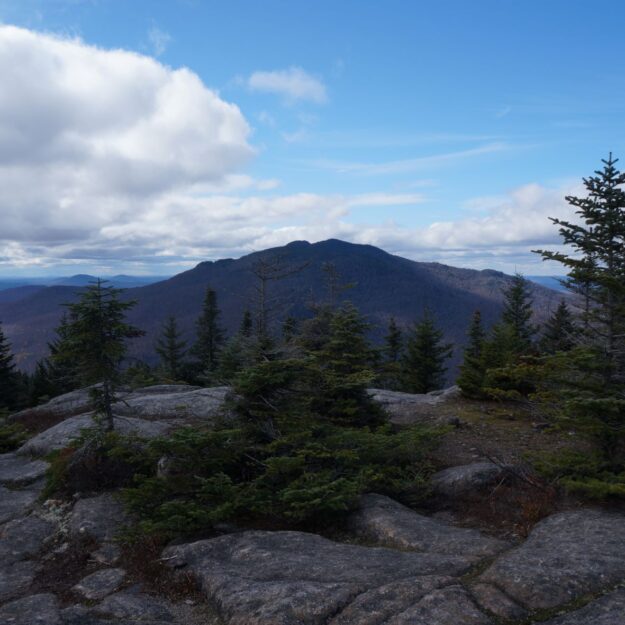 Rock face mountain summit looking toward alpine trees and another small mountain in distance