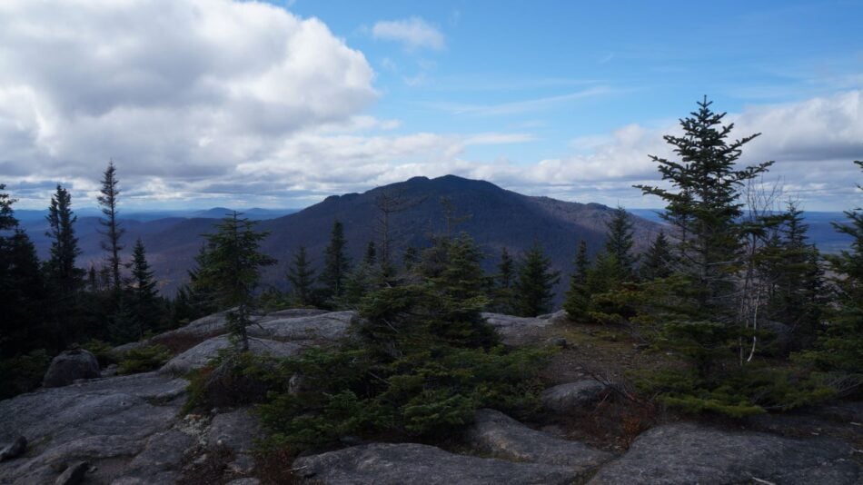 Rock face mountain summit looking toward alpine trees and another small mountain in distance