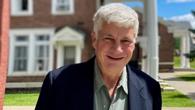 Man with white hair wearing a navy blue blazer and light green button down shirt stands in front of a county building with red bricks and white trim