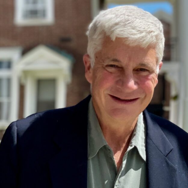Man with white hair wearing a navy blue blazer and light green button down shirt stands in front of a county building with red bricks and white trim