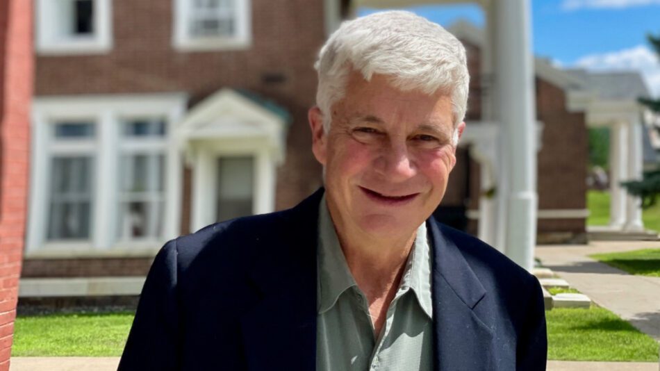 Man with white hair wearing a navy blue blazer and light green button down shirt stands in front of a county building with red bricks and white trim