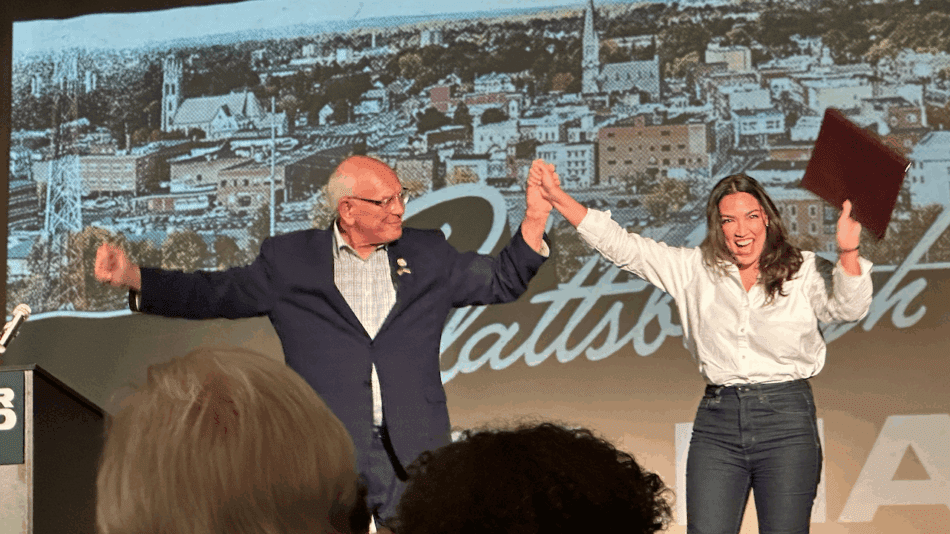 A man and a woman hold hands on stage and cheer to a crowd.