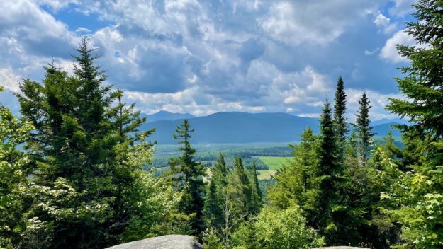 Summit vista looking out from side of mountain rock surface toward alpine trees and Adirondack mountains in the distance.