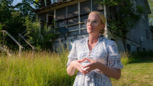 A blonde woman with glasses in a white dress standing in front of an overgrown grass/weeds with an old house behind it.