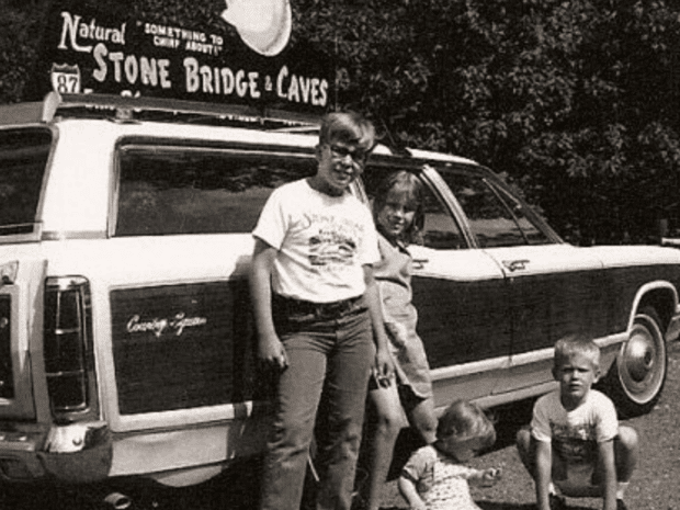 Black and white image of young kids and toddler in front of a station wage with a sign on top that says Natural Stone Bridge & Caves