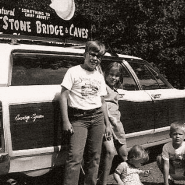 Black and white image of young kids and toddler in front of a station wage with a sign on top that says Natural Stone Bridge & Caves