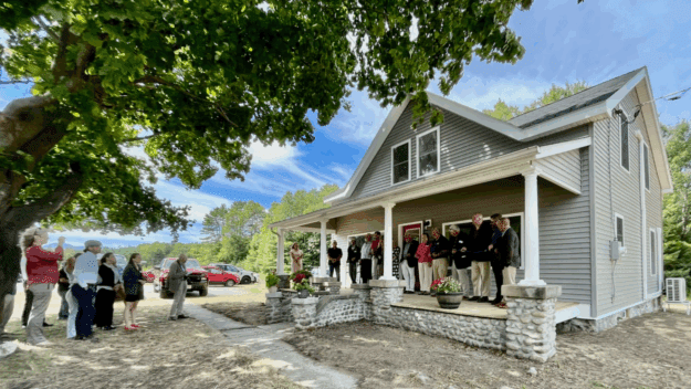 People line up in front of an small gray farmhouse with white columns on stone pillars
