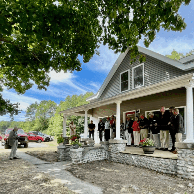 People line up in front of an small gray farmhouse with white columns on stone pillars