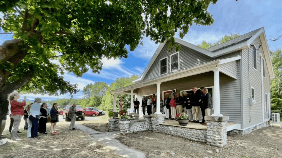 People line up in front of an small gray farmhouse with white columns on stone pillars