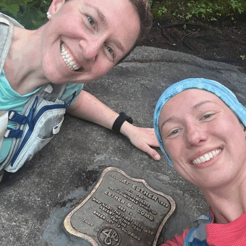 Debbie McElwaine and Katie Rhodes on Mt. Esther posing on the ground close up with the plaque on Mt. Esther