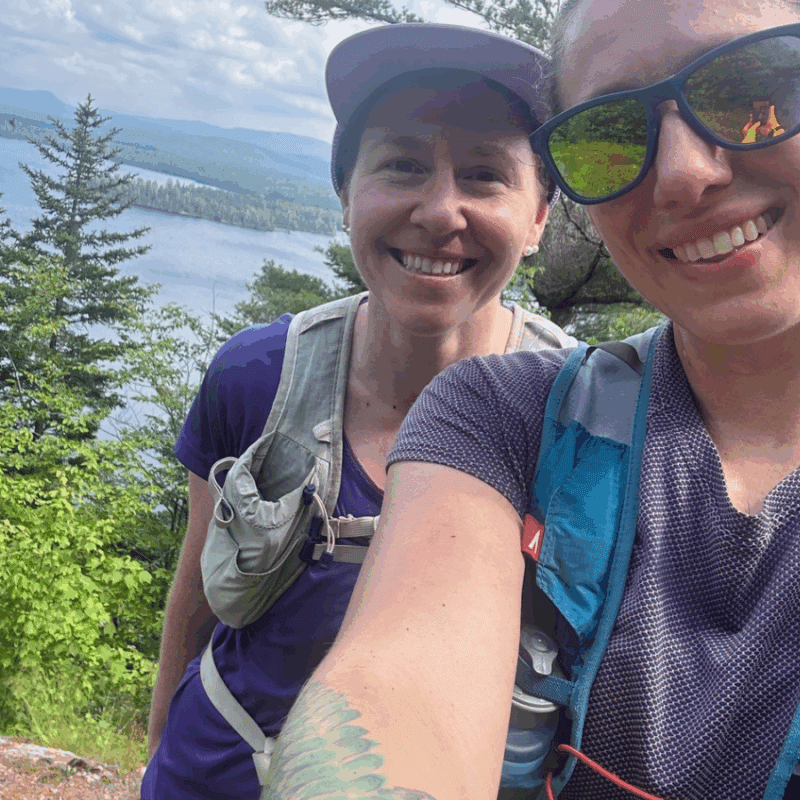 Selfie photo of Debbie McElwaine and Katie Rhodes on Frederica Mountain with evergreen trees and lake views in distance.